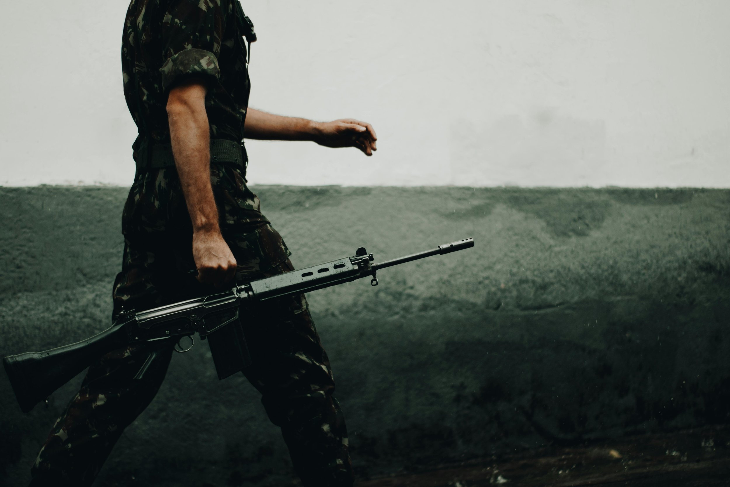 A person wearing military camouflage clothing holding a rifle in their right hand, walking against a plain background.