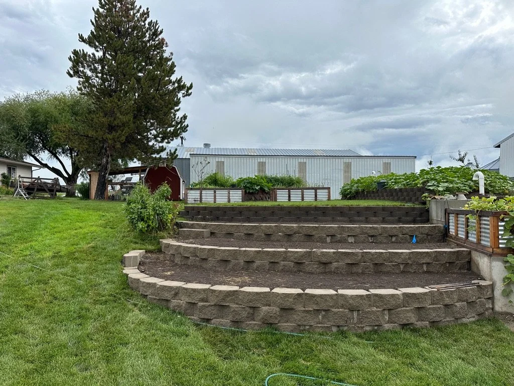 A backyard with stone steps leading up to a garden area with raised beds, trees, and a shed, on a cloudy day.