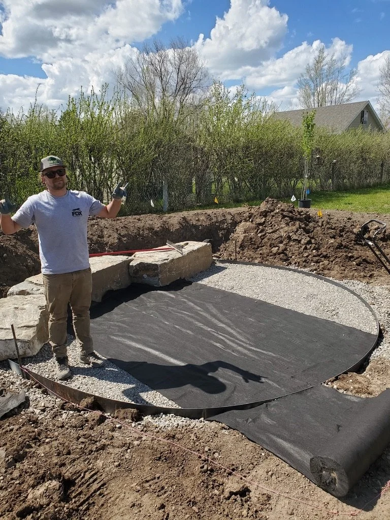 A man standing in a backyard construction site, giving a thumbs-up near a circular area being prepared with landscape fabric and border edging, with large rocks and gravel around.