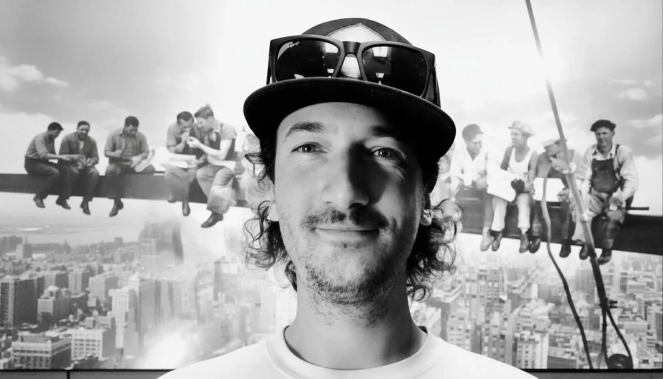 A man taking a selfie in front of a black-and-white mural of the 'Lunch atop a Skyscraper' photo, with construction workers sitting on a steel beam.