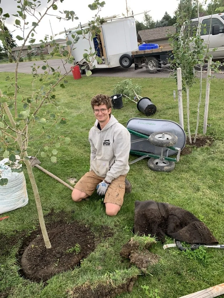 A young man kneeling on the grass next to a newly planted small tree, holding a gardening tool. There are gardening supplies, a wheelbarrow, and a young tree in a burlap sack nearby. In the background, there is a trailer, a parked van, and a few trees.