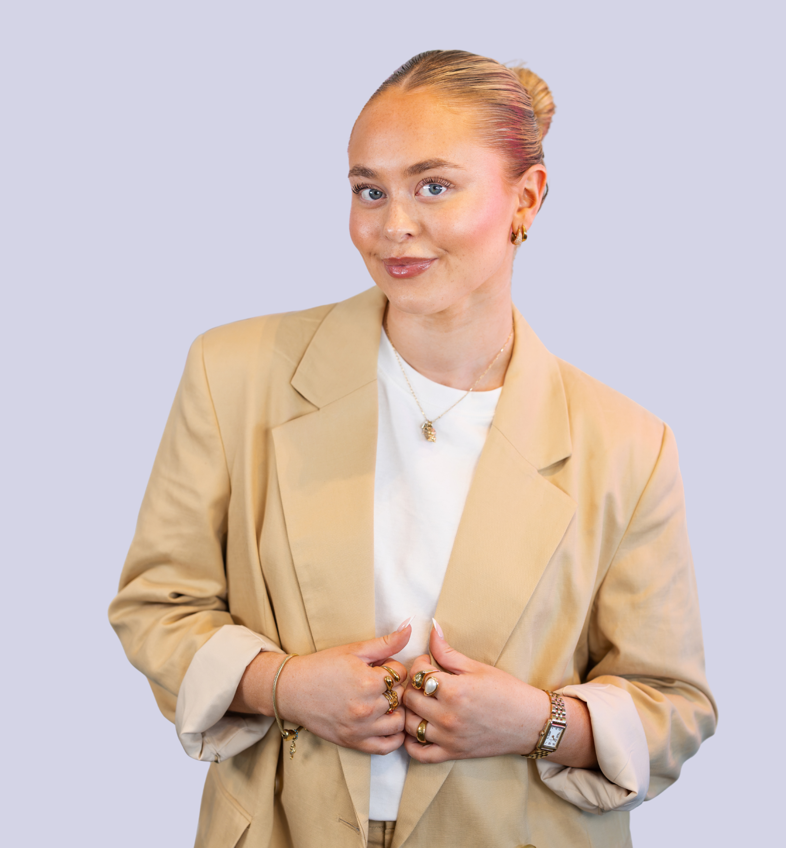 Kvinde med blondagt hår i hestehale, iført beige blazer og hvid T-skjorte, smiler. Bærer ringe, ur og smykker, professionel baggrund.