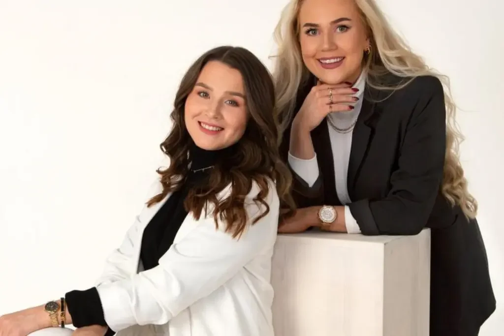 To kvinner, en med mørkt hår i en hvit jakke og en med blond hår i svart dress, smiler mot kamera.