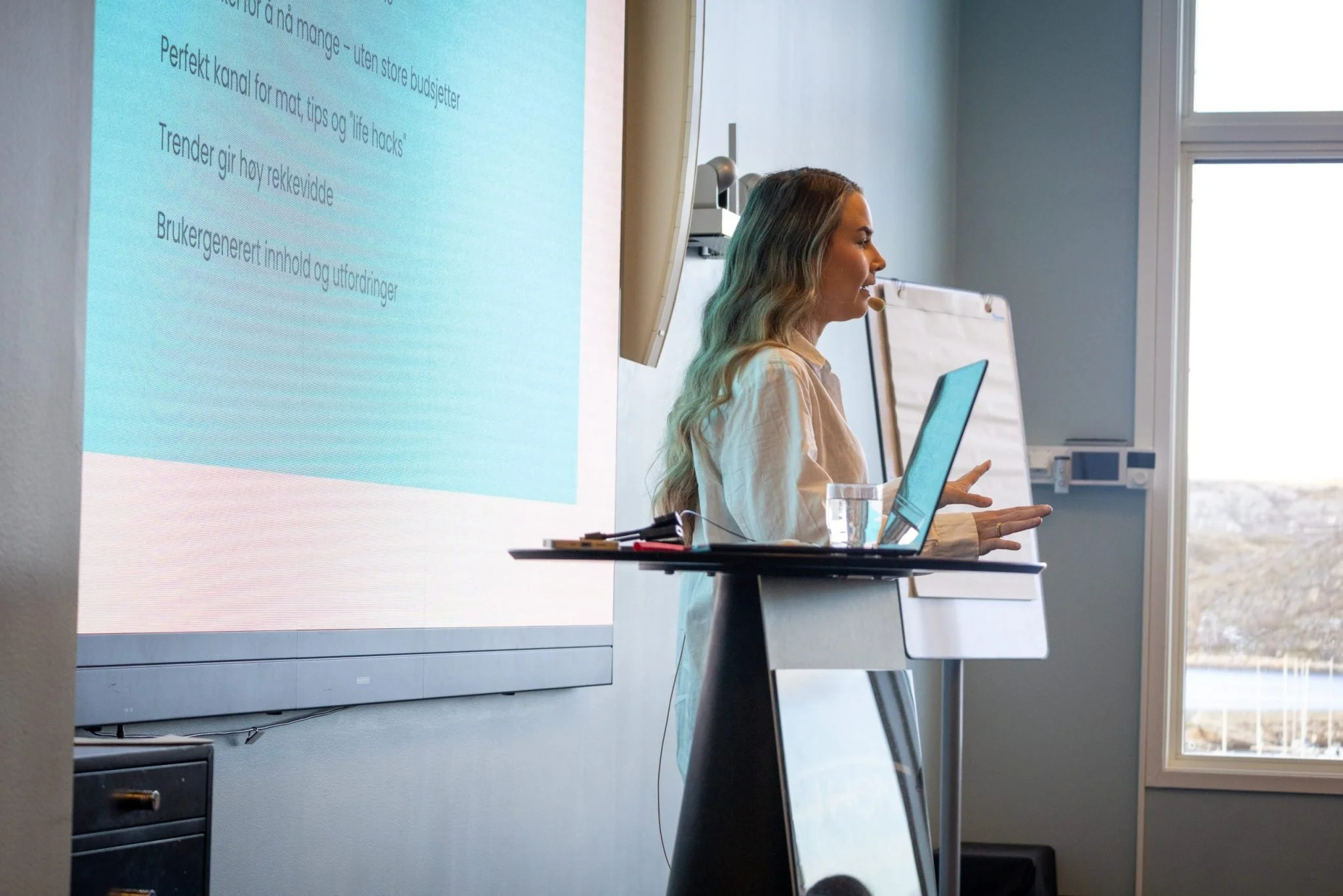 En kvinne holder en mikrofon og sitter foran en laptop i et møterom, med en projeksjon på veggen som viser tekst på norsk.