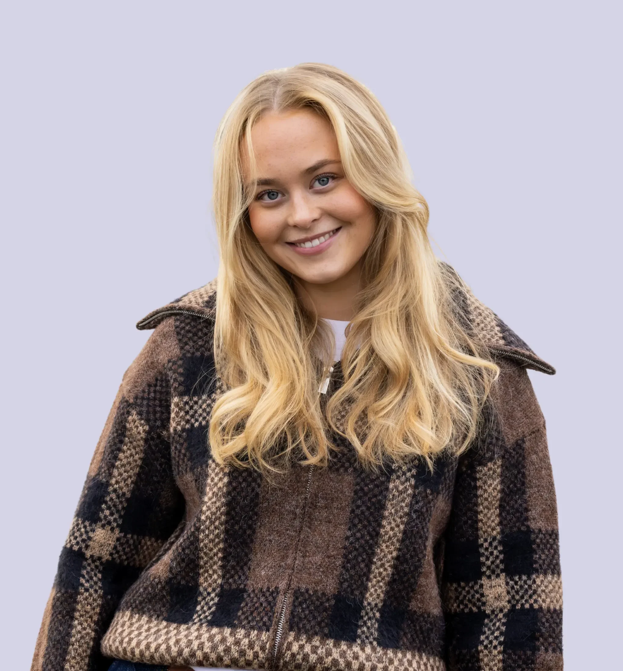 Et smilende ungt kvinne med langt blondt hår, iført en brun og svart rutete pelsjakke, står mot en lys lilla bakgrunn.