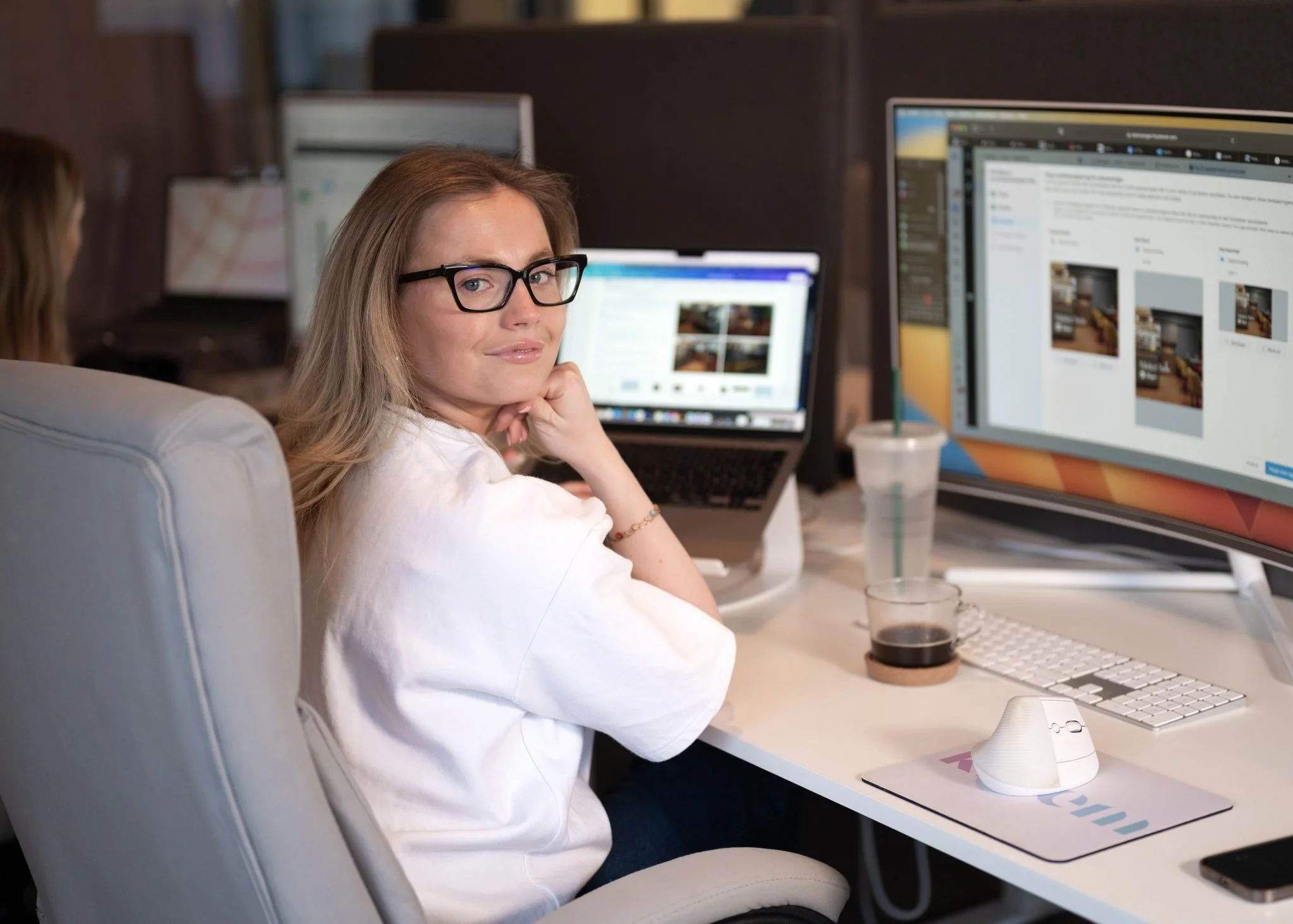 Kvinne med briller sitter ved datamaskin på kontor, omgitt av flere skjermer, og glaner mot kameraet.