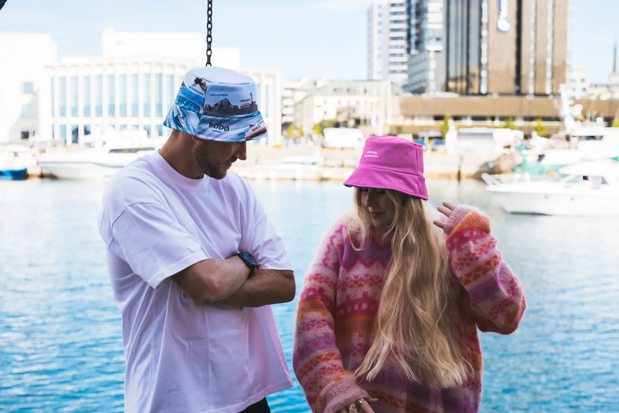 To personer står ved vannet, en mann og en kvinne, begge har på seg fargerike, brede hatter. Mannen har hvitt t-skjorte og kvinnens plagg er rosa og oransje. Bak dem er det flere båter og høye bygninger i en by.