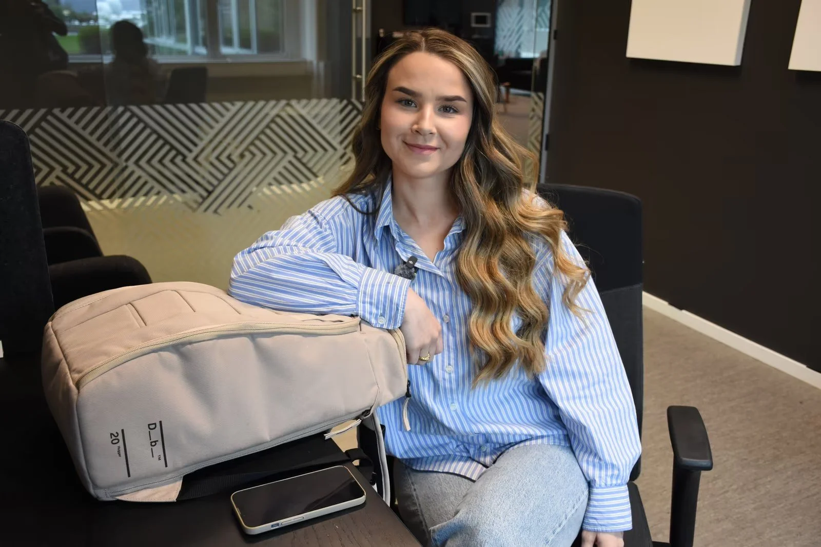 Kvinne med langt, bølget hår og i blåstripet skjorte sitter ved et bord i et kontor, med en beige ryggsekk og en smarttelefon foran seg.