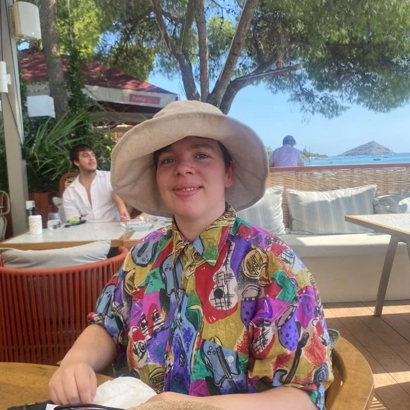 A young boy smiling at the camera, wearing a colorful patterned shirt and a large beige sun hat, sitting at a table in an outdoor restaurant with a view of the beach and ocean in the background.