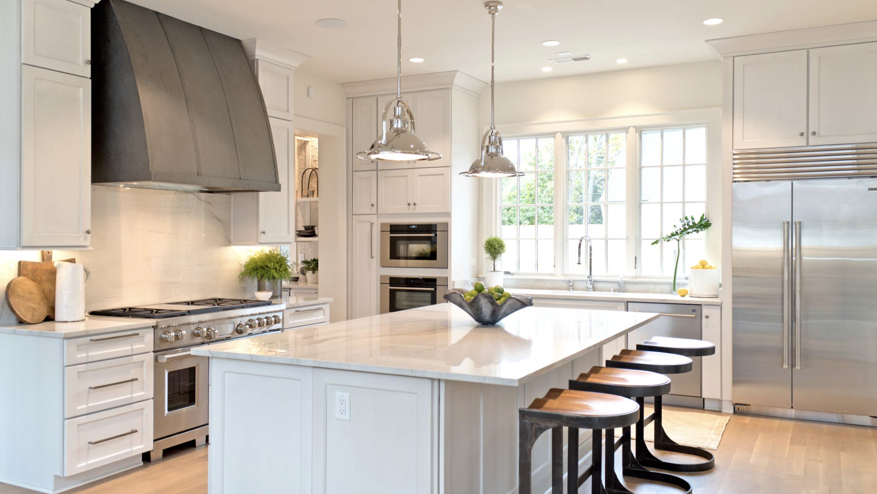 Bright, modern kitchen with white cabinets, stainless steel appliances, a large island with a white marble countertop, black wooden stools, and a window with greenery outside.