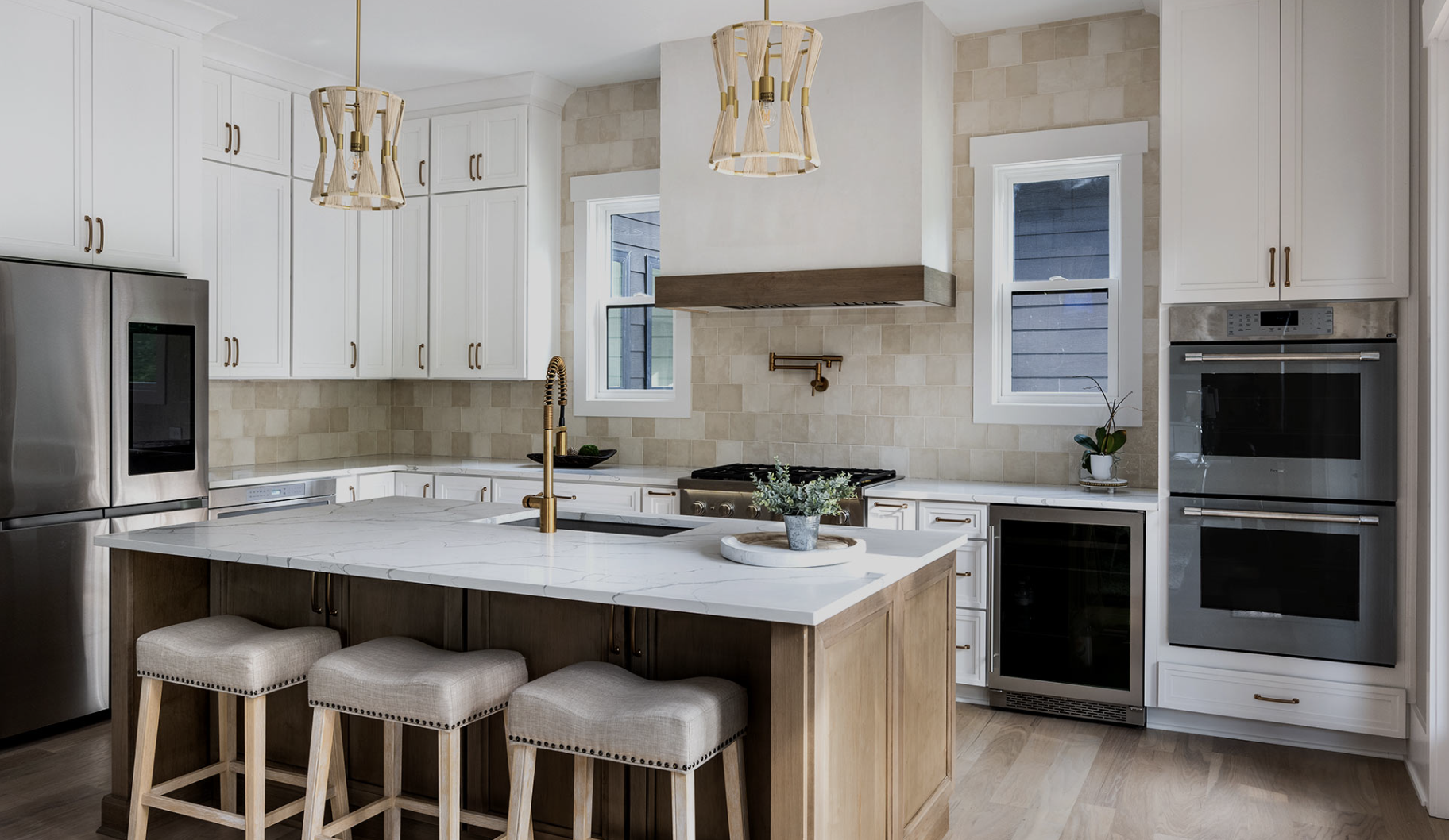 Modern kitchen with white cabinets, beige tile backsplash, kitchen island with marble countertop, beige bar stools, stainless steel appliances, pendant lights, small potted plant, and windows with white trim.