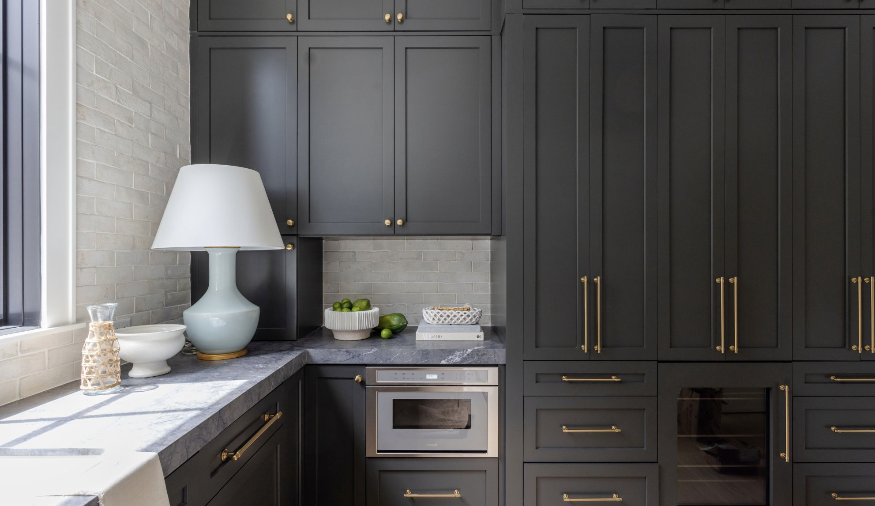 Kitchen with dark gray cabinets, marble countertop, gray brick backsplash, white table lamp, decorative bowls, and green limes.