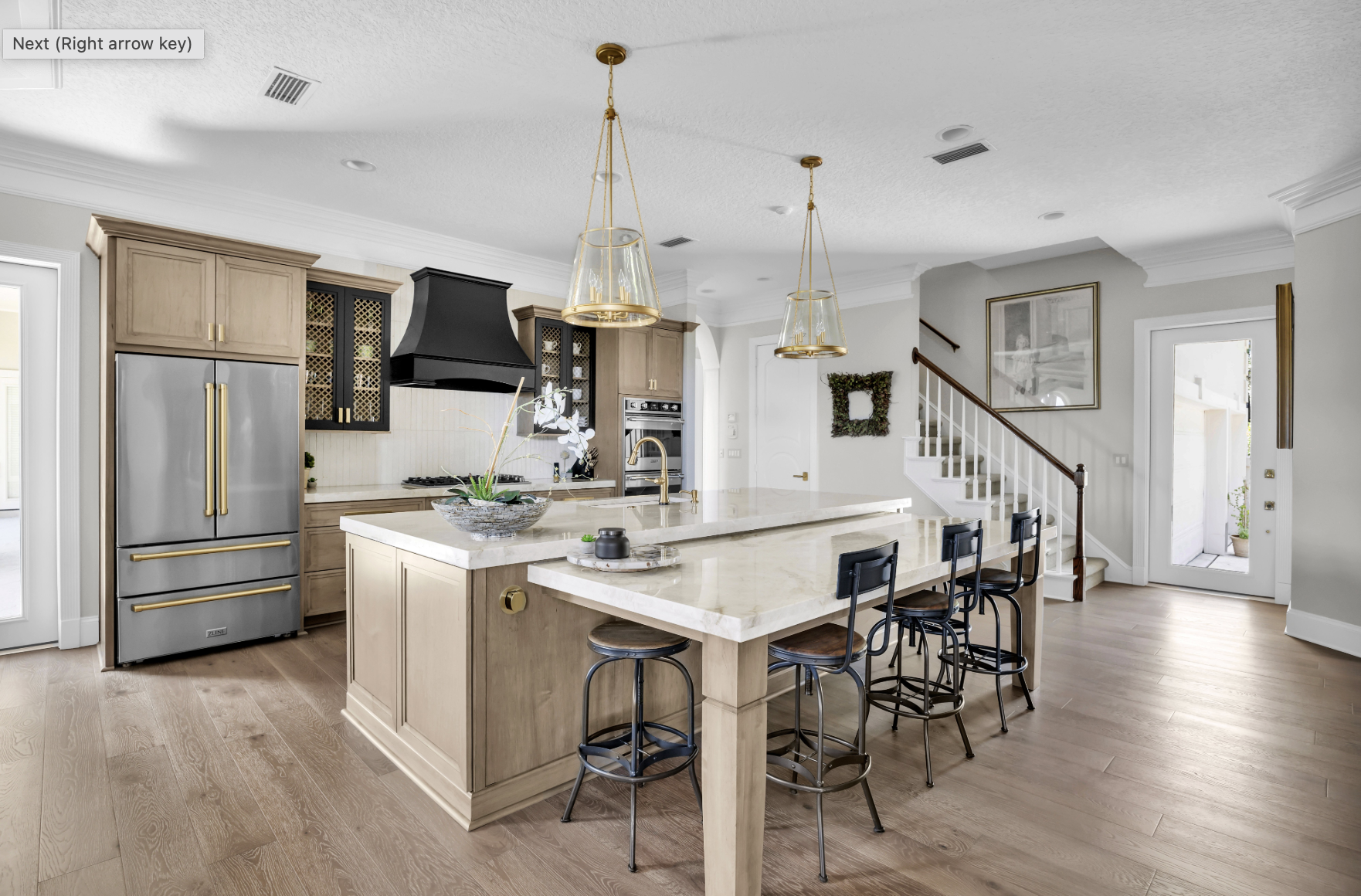 Modern kitchen with beige cabinets, stainless steel appliances, and a large island with bar seating. Pendant lights hang over the island. There is a staircase leading to an upper floor near a door that opens to an outdoor area.