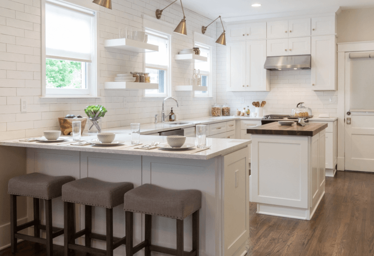 Bright white kitchen with three stools at a breakfast bar, open shelves with dishes, windows letting in natural light, and a granite countertop.