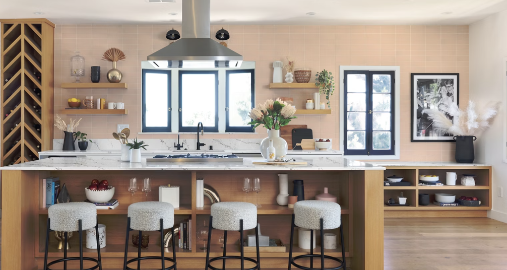 Modern kitchen with pink tiled wall, island with barstools, open shelving, and decorations.