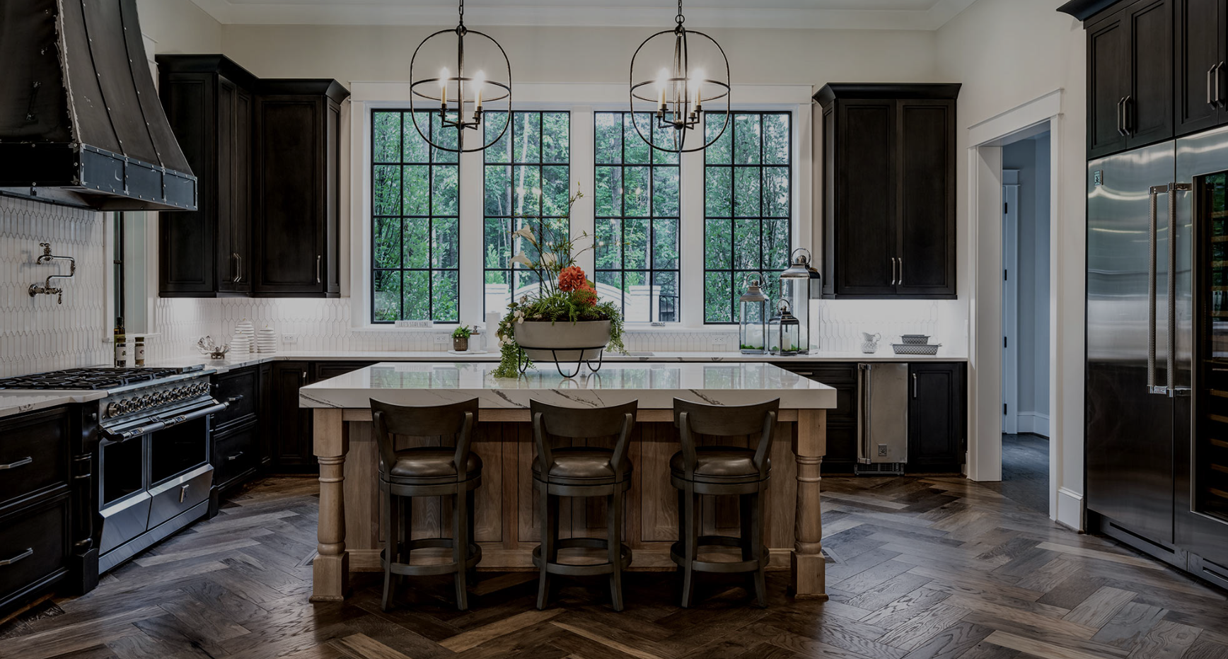Modern kitchen with black cabinets, white countertops, a central island with three chairs, large window with green trees outside, and two hanging light fixtures.