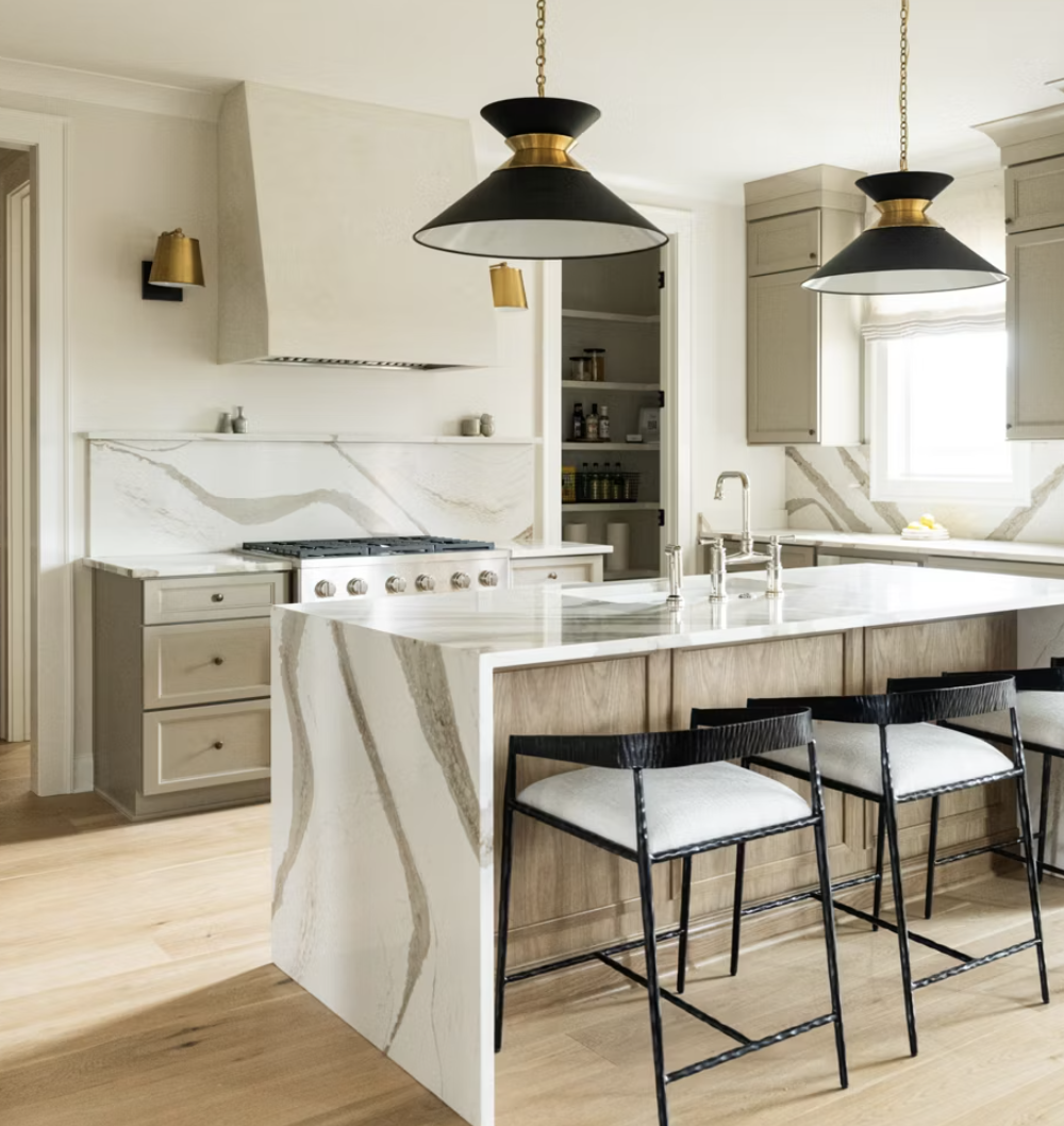 Modern kitchen with white marble countertops and backsplash, black and gold pendant lights, wooden bar stools with white cushions, and light cabinetry.