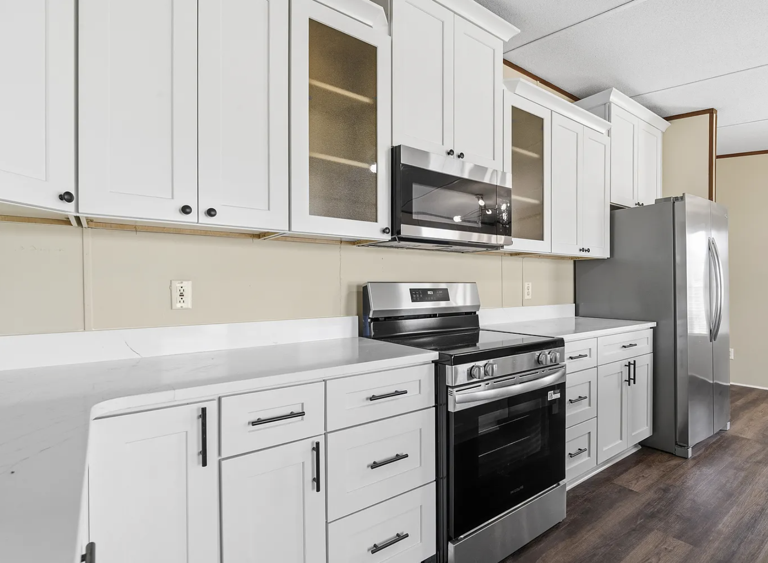 Kitchen with white cabinets, stainless steel refrigerator, microwave, and oven on dark brown wooden floor, beige wall, and ceiling with brown trim.