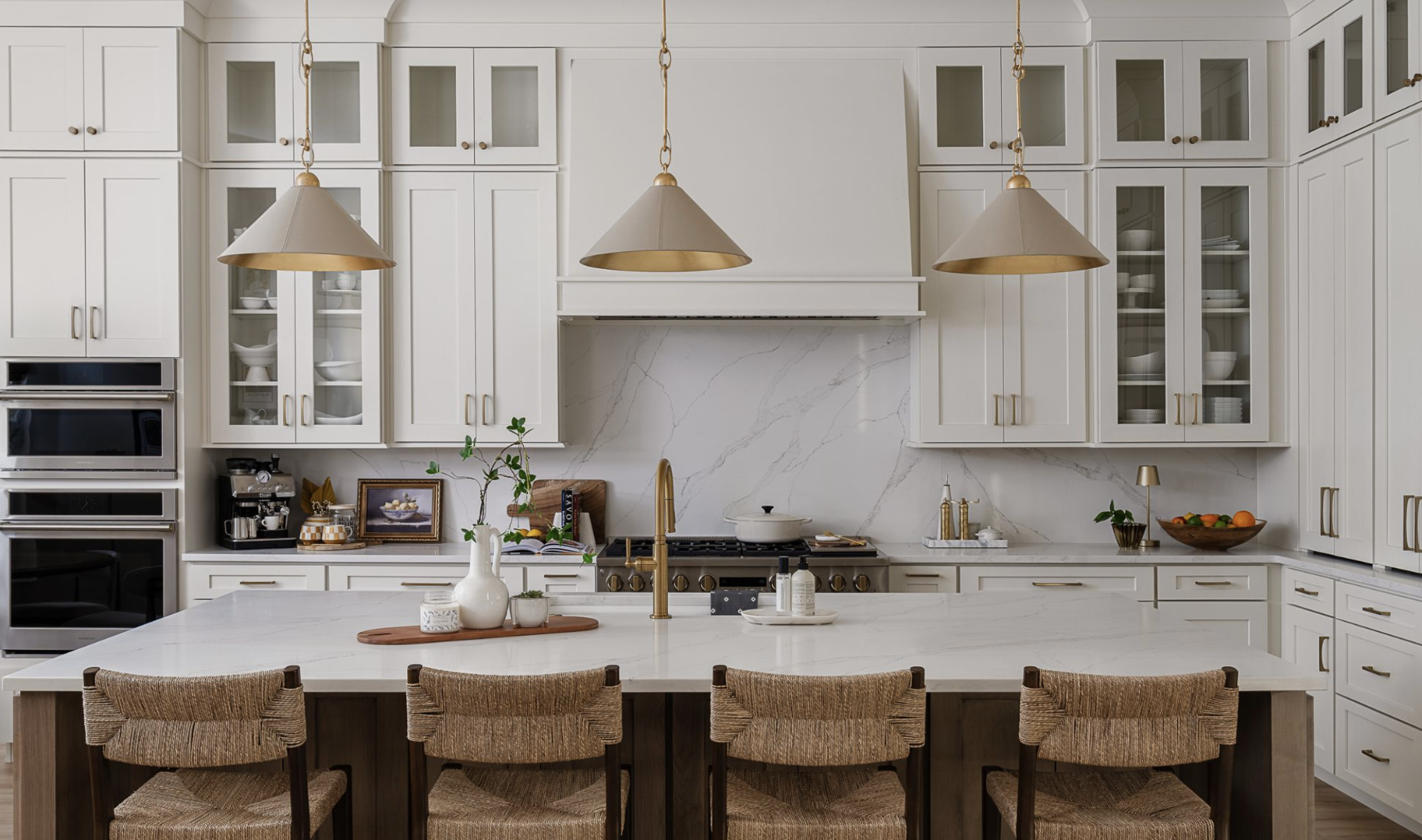 Modern kitchen with white cabinetry, gold hardware, and pendant lights over a large island with wicker chairs. White marble backsplash and countertops, with various kitchen accessories, plants, and bowls of fruit.