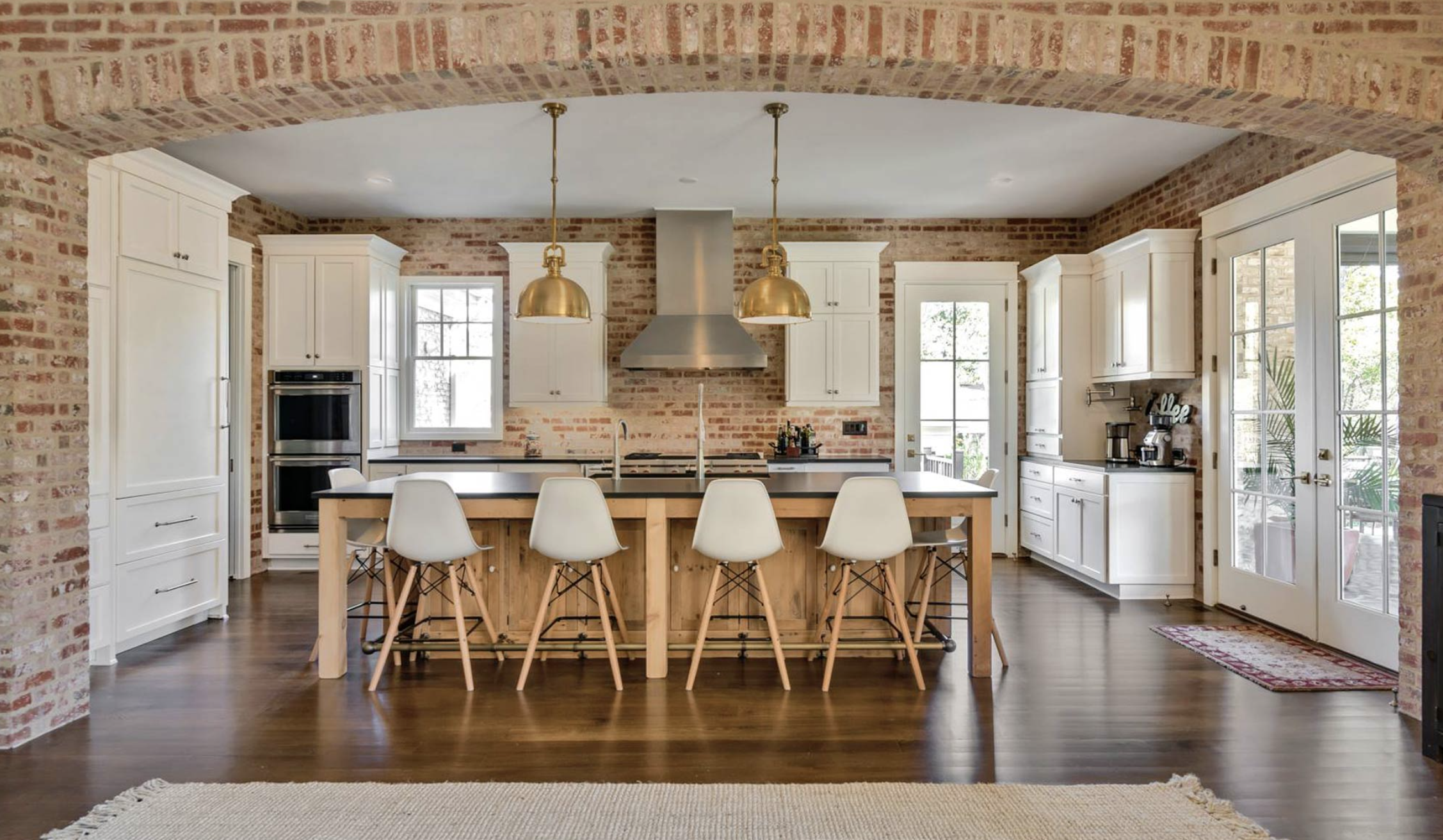 Kitchen with exposed brick walls, white cabinets, a central island with a wooden base, six white chairs, pendant lights, and multiple windows.