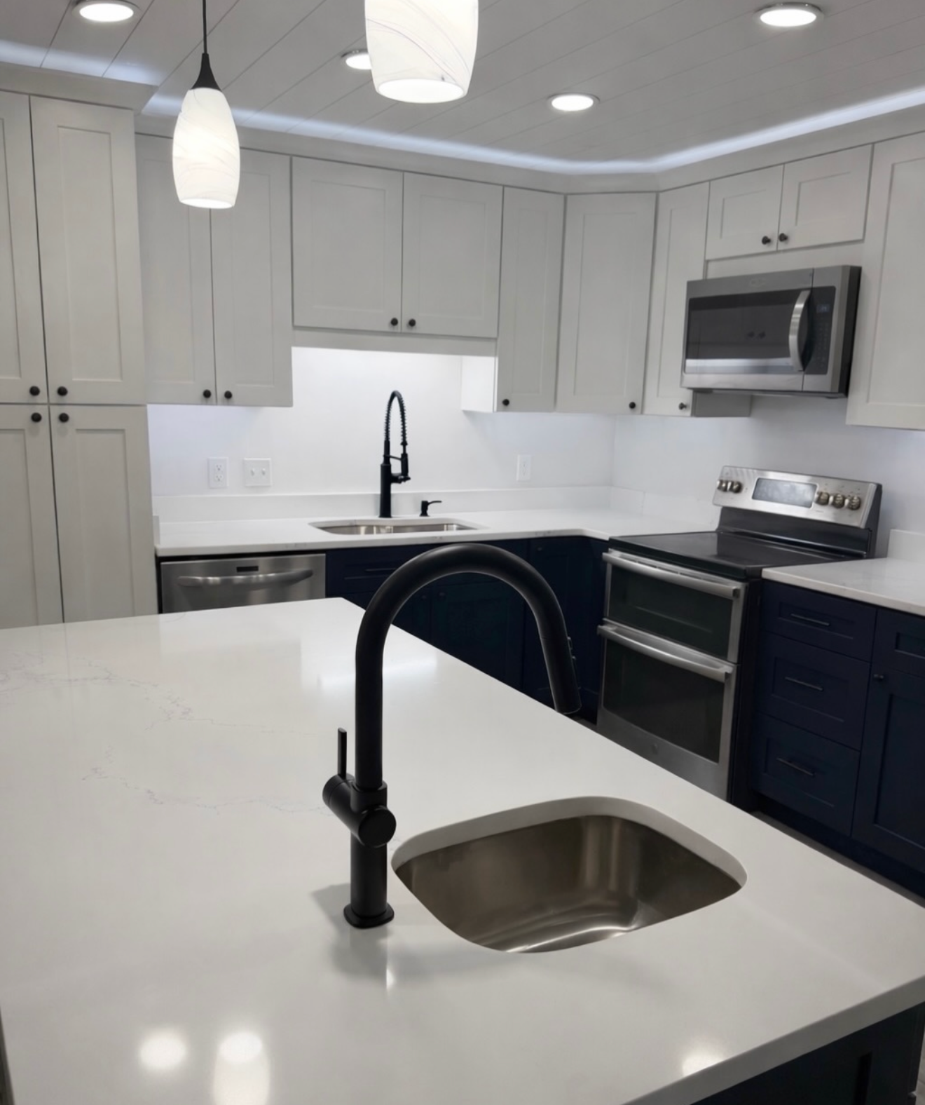 A modern kitchen with white and dark blue cabinets, a white countertop, a black faucet, stainless steel appliances including a microwave and oven, and a black sink. There are ceiling lights and pendant lights.