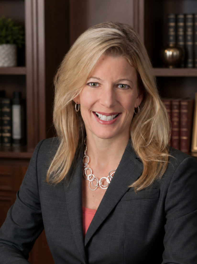 A smiling middle-aged woman with blonde hair, wearing a dark suit jacket and a necklace, sitting at a desk in an office with wooden bookshelves in the background.