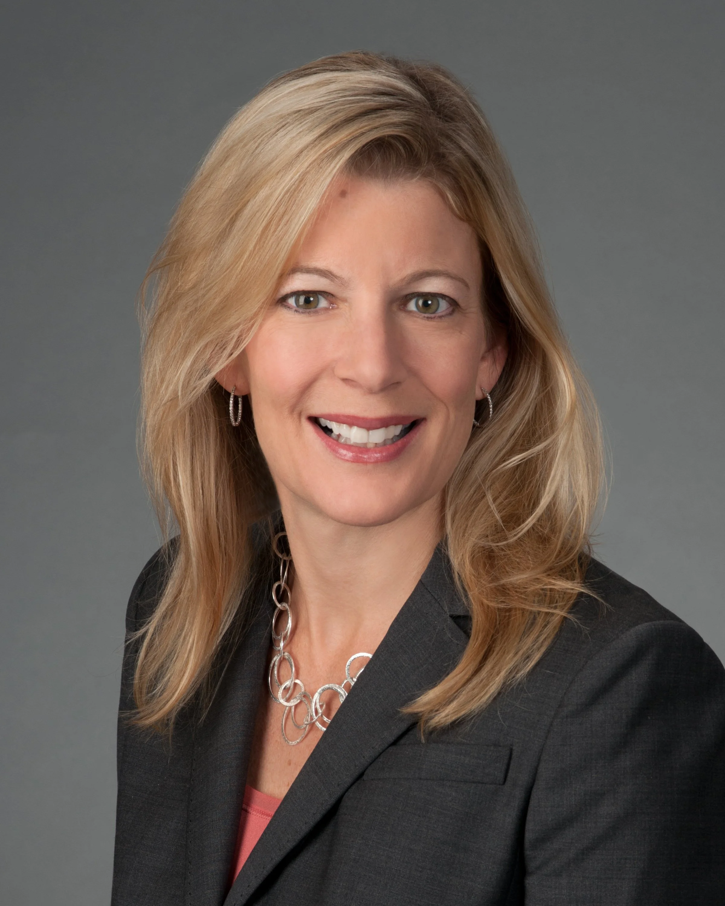 A woman with blonde hair wearing a dark blazer, pink top, silver earrings, and a silver chain necklace, smiling against a gray background.