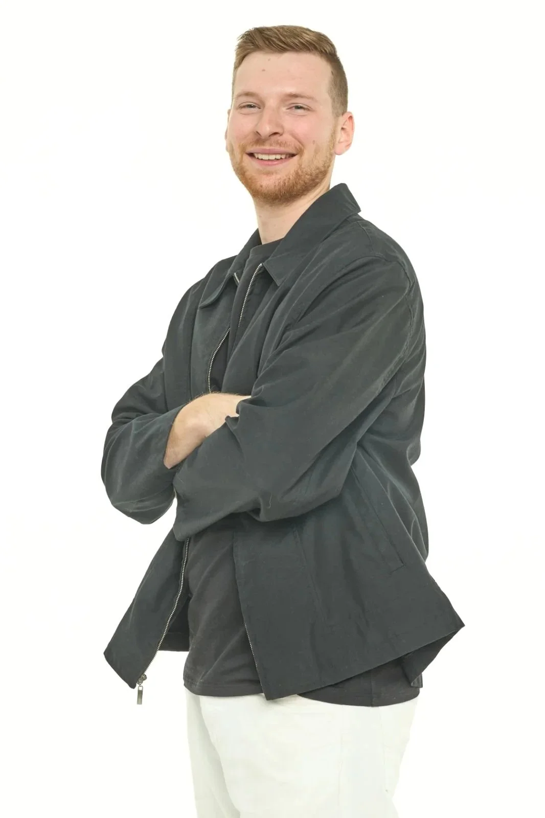 Un homme souriant avec une barbe, portant une veste noire et un pantalon blanc, avec les bras croisés, isolé sur fond blanc.