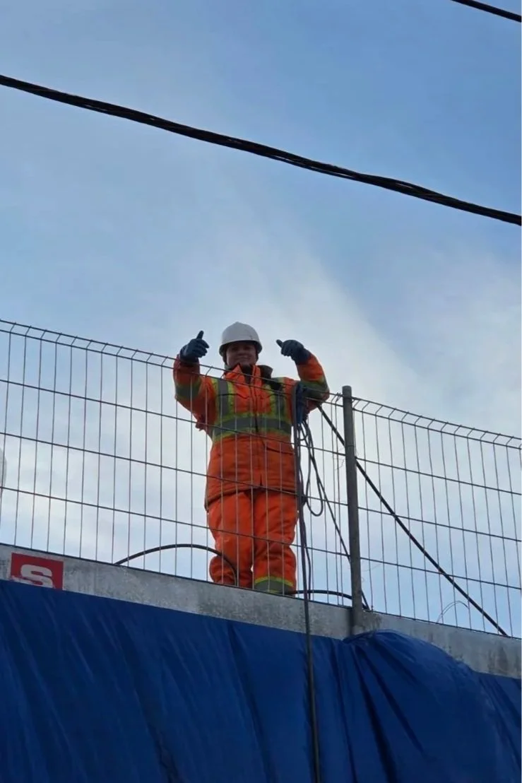 Un ouvrier en tenue de sécurité orange et casque blanc, debout sur une plateforme en hauteur, donne un pouce en l'air. Il semble travailler sur une structure ou un chantier avec un ciel clair en arrière-plan.