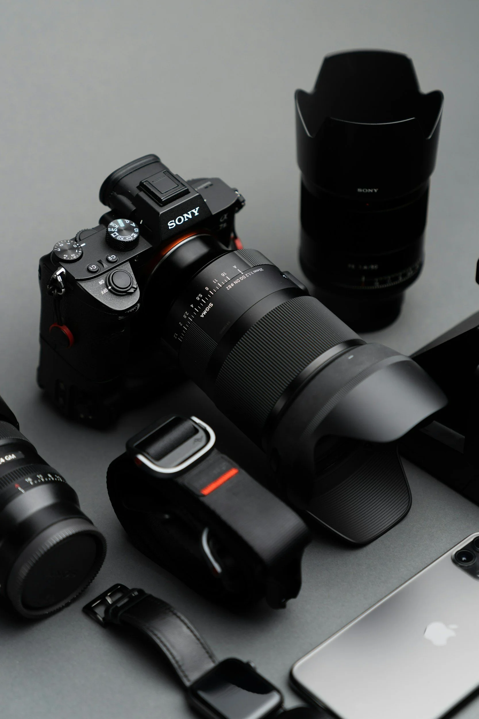 A collection of photography equipment including a Sony camera with a large lens, a second lens, a camera strap, a flashlight, a smartphone, and other camera accessories on a grey surface.