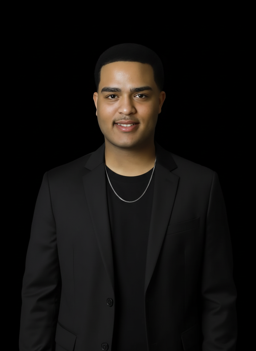 Portrait of a young man with short black hair, wearing a black blazer over a black shirt, and a silver chain necklace, smiling against a black background.