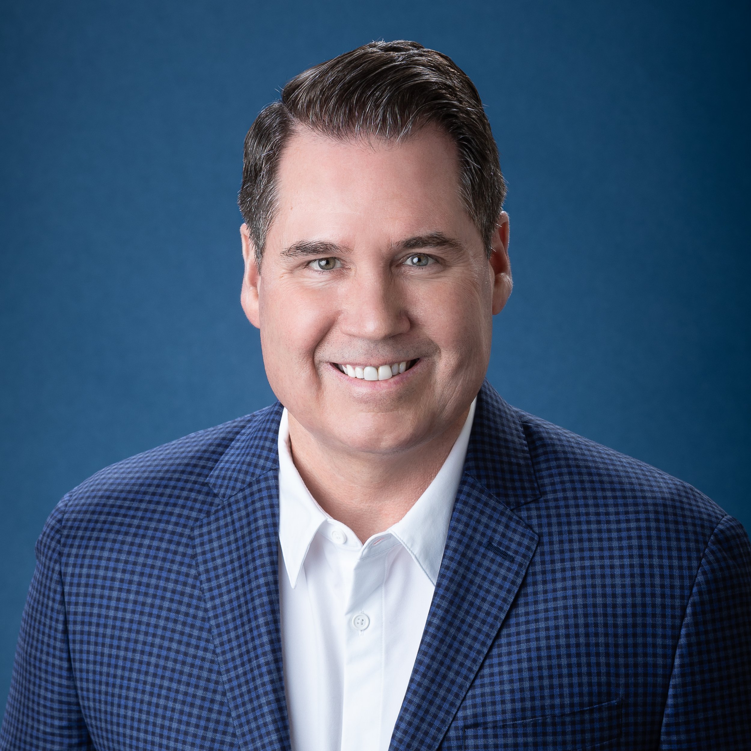 Professional headshot of a smiling man with dark hair in a checkered blue blazer and white shirt, against a solid blue background.