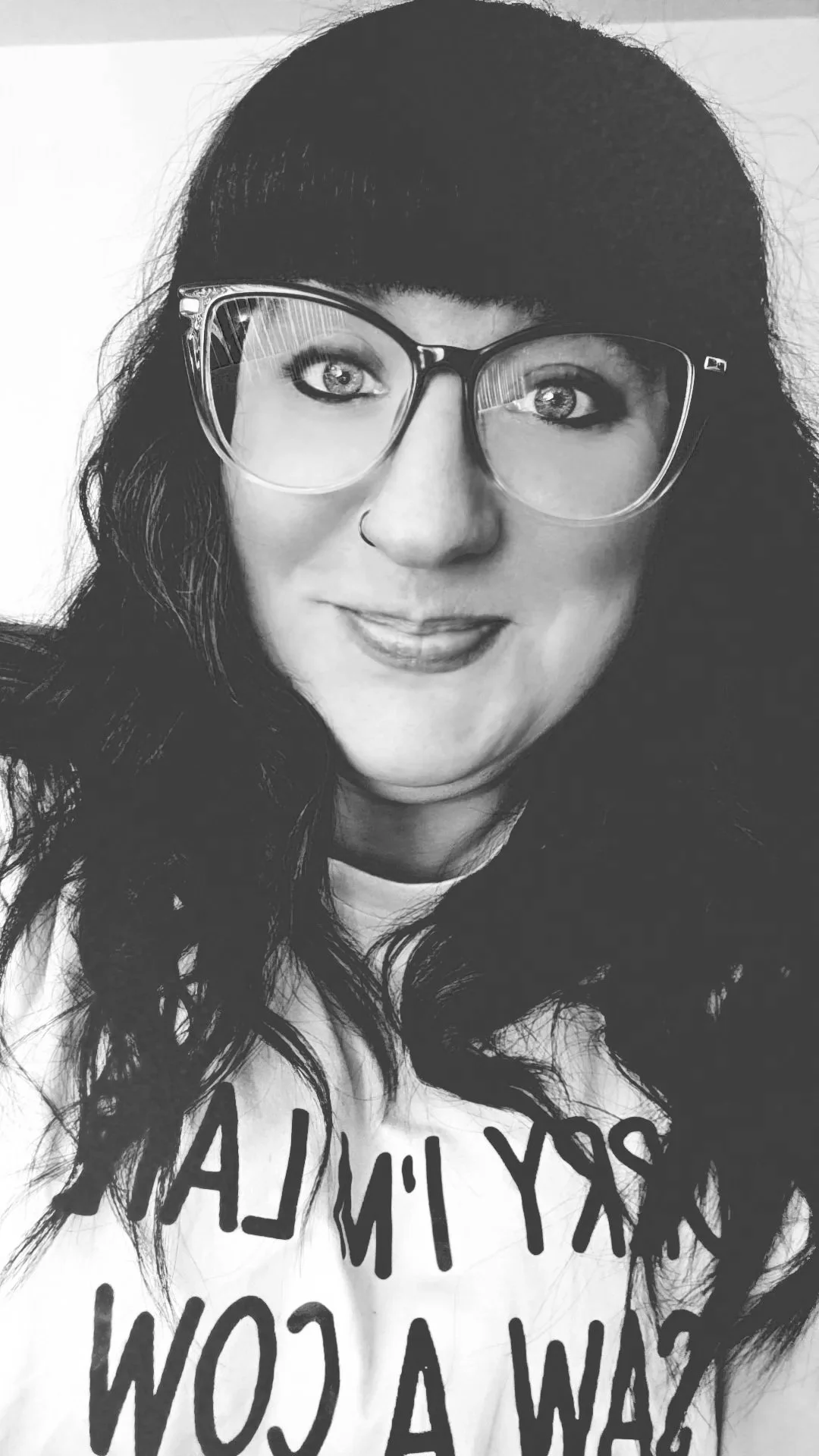 Black and white photo of a woman with long wavy hair, wearing large eyeglasses, a nose ring, and a T-shirt with partly visible text.
