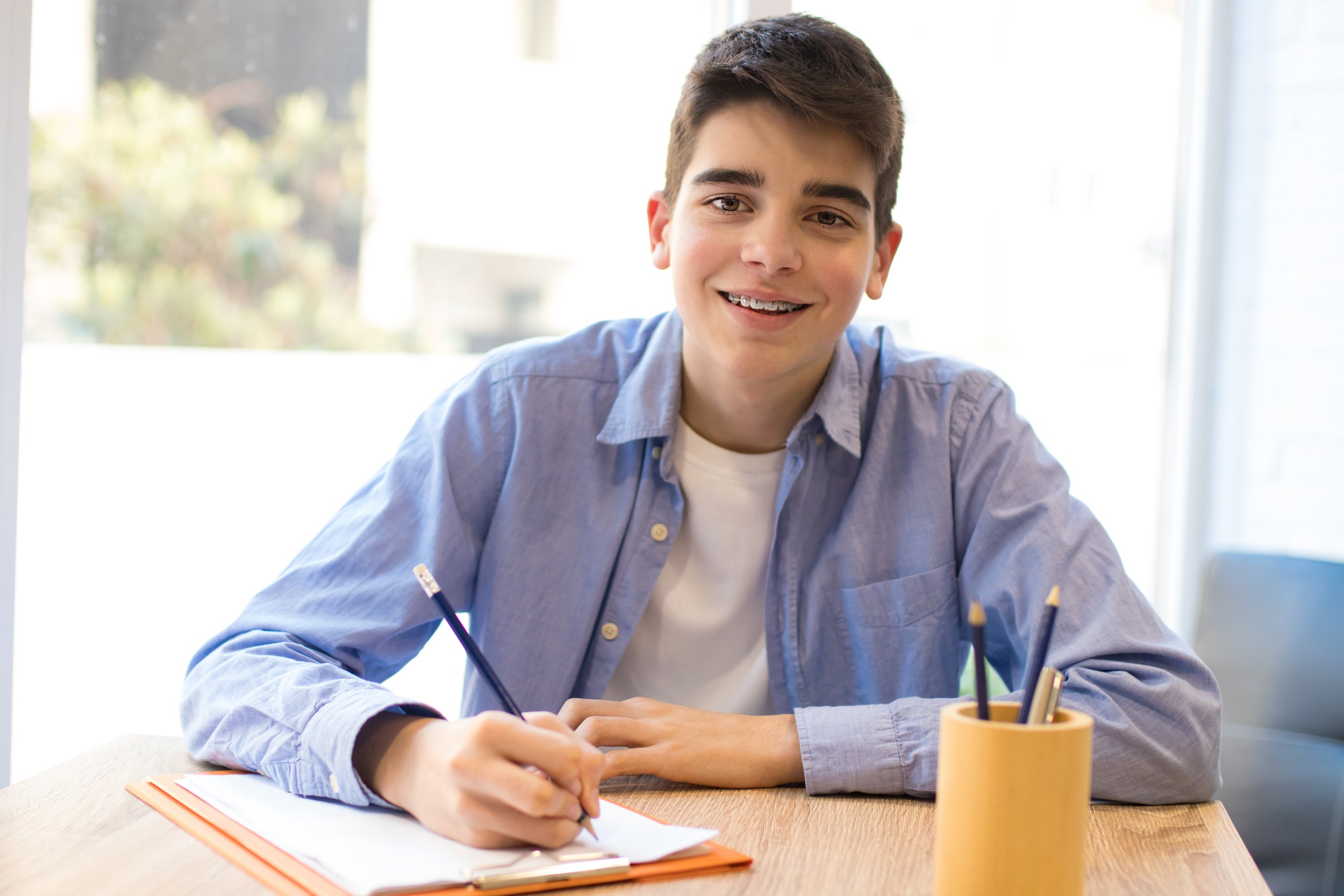 A teenage boy with dark hair smiling, sitting at a desk with a notebook and pens, in a room with large windows and natural light.
