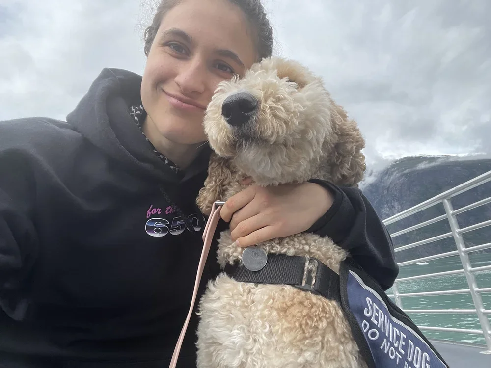 A woman smiling and holding a service dog with a harness labeled 'SERVICE DOG DO NOT PET' on a boat or dock with water and mountains in the background.