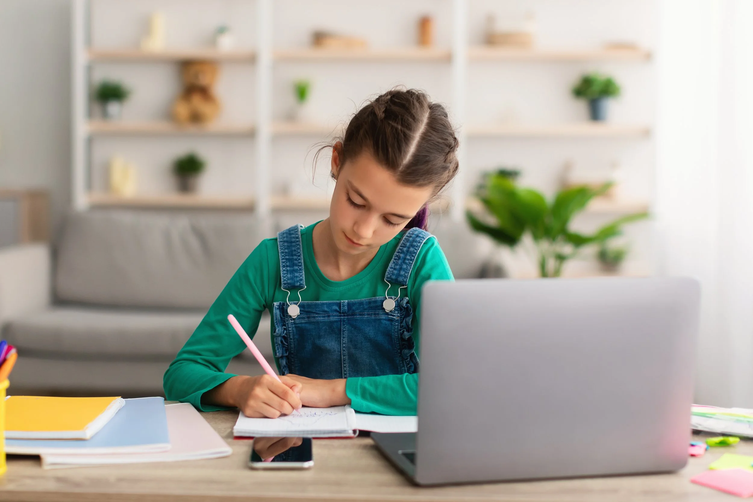 Young girl with braided hair wearing green shirt and denim overalls sitting at a desk, writing in a notebook with a pink pen, a laptop, and colorful stationery on the desk, in a bright room with shelves and plants in the background.