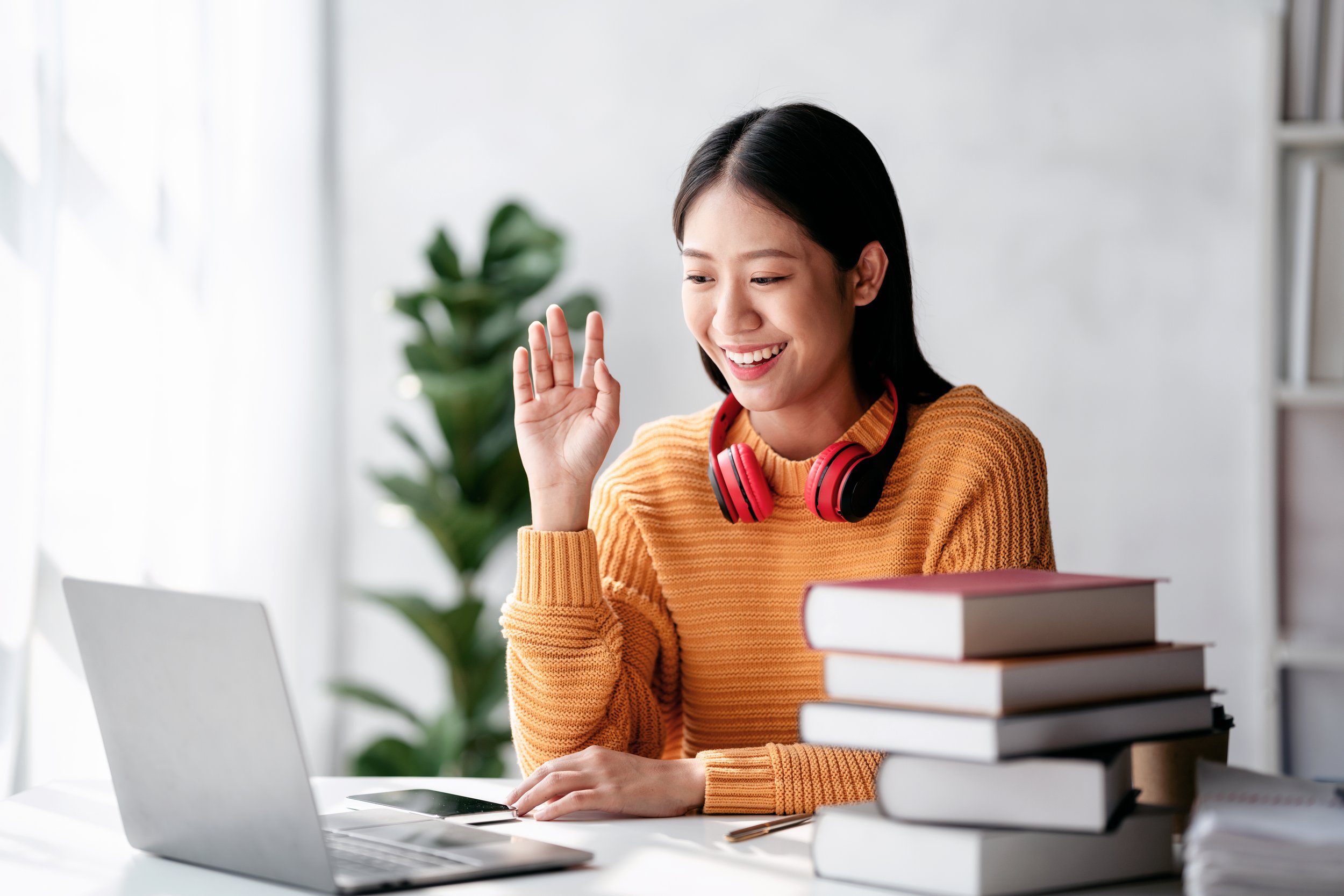 A woman with headphones smiling and waving at her laptop in a bright room with a potted plant in the background.