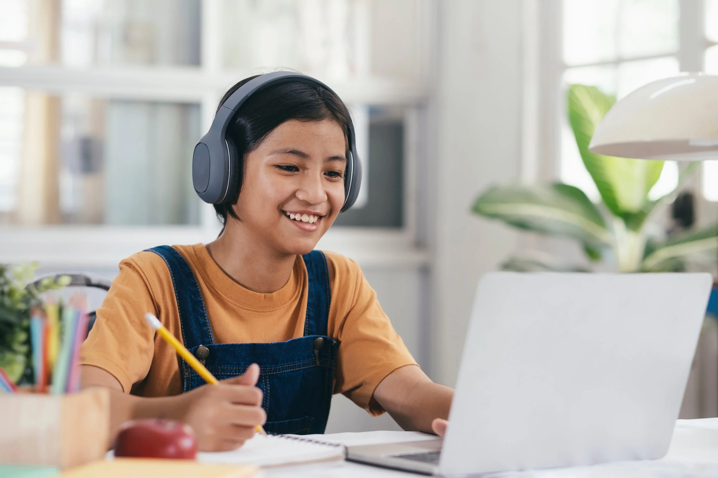 A young girl with short black hair, wearing headphones, a yellow shirt, and denim overalls, is smiling while working on a laptop at a desk with a notebook and a yellow pencil in her hand.