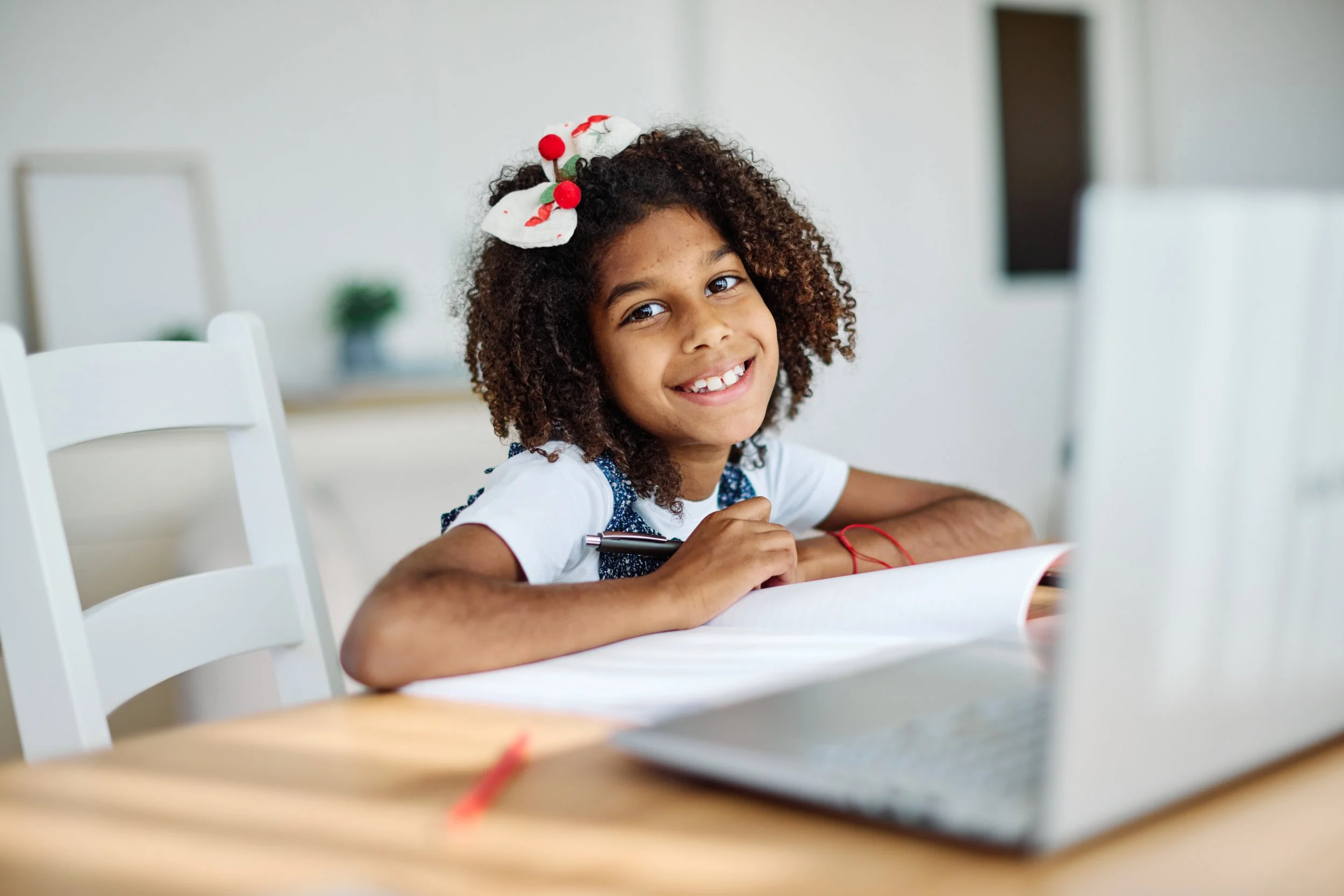 A young girl with curly hair, smiling, wearing a holiday-themed bow, sitting at a table with a notebook and pen, looking at a laptop.