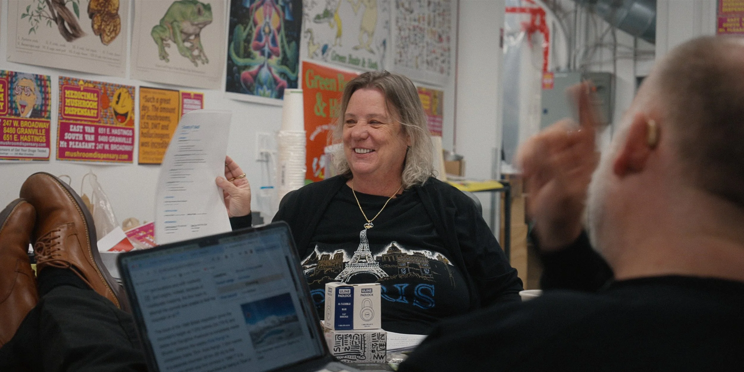 Cindy Heemeryck, the manager of the Coca Leaf Cafe is shown in this image. Cindy is smiling while holding a piece of paper, seated at a table with two men using laptops, in a room decorated with colourful drug posters and artwork on the walls.