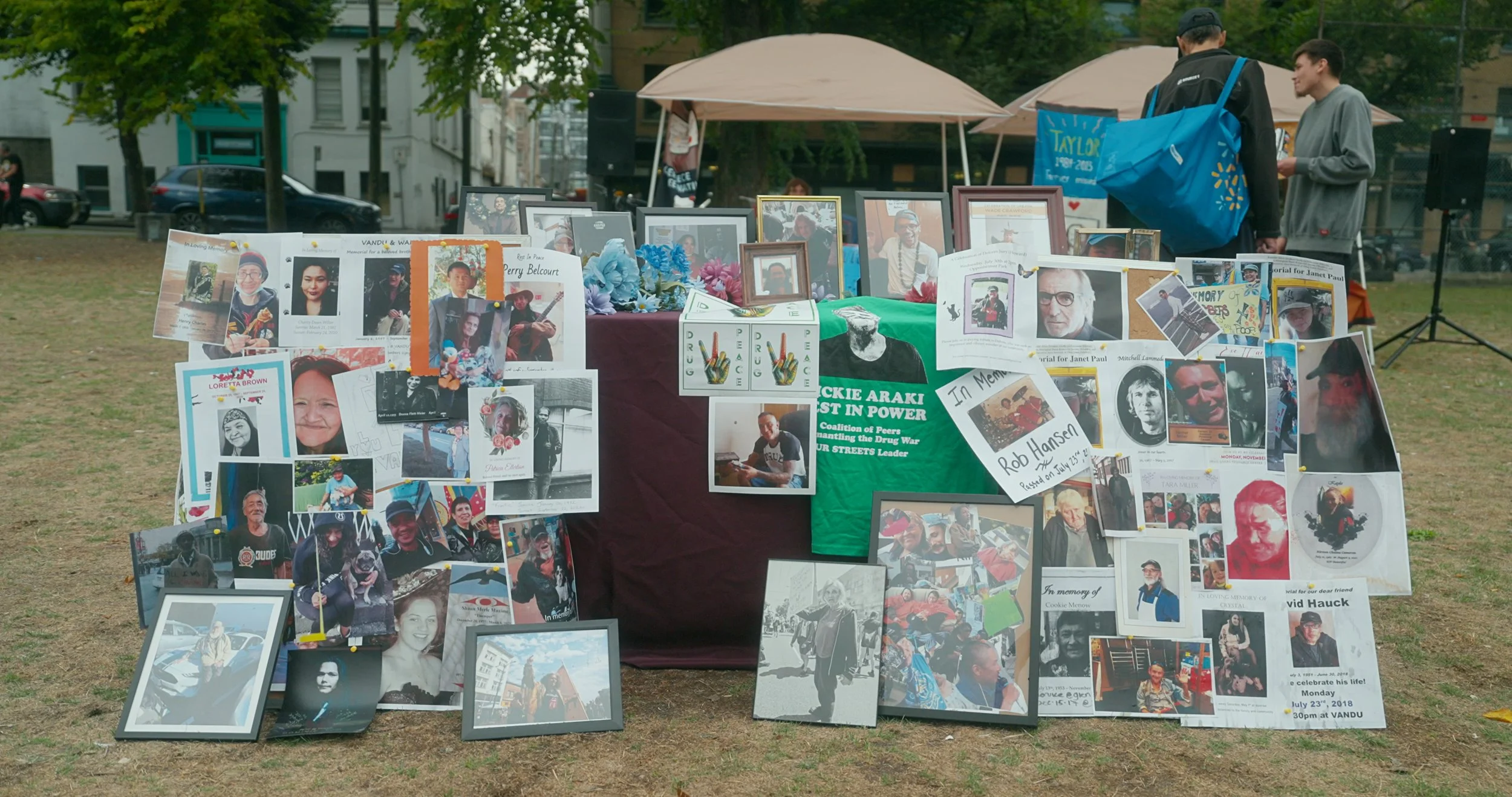 A memorial display of the lives lost to the Toxic Drug crisis. Shown are numerous photographs, posters, and signs honoring these lives. The setup is outdoors, with tables, umbrellas, and trees in the background, and people observing the memorial.