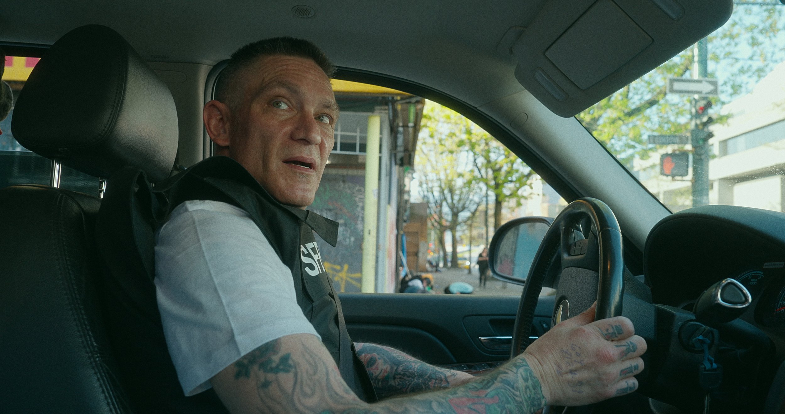 Jerry Martin, a drug activist based in Vancouver is driving to open up The Drugs Store, an illegal store selling hard drugs. He is wearing a white shirt and vest, sitting in the driver's seat of a car, looking back at camera with a slight smile.