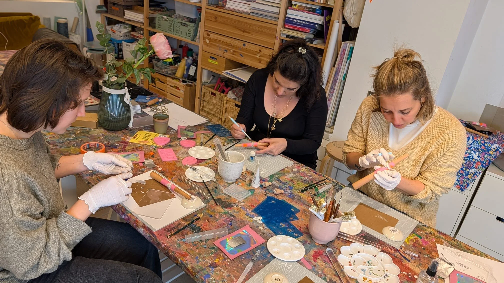 Drei Frauen sitzen an einem bunten, verschmutzten Tisch, arbeiten an bunten, reflektierenden Gegenständen, vermutlich Schmuck oder Kunsthandwerk, in einem Raum mit Regalen voller Bücher und Bastelmaterialien.