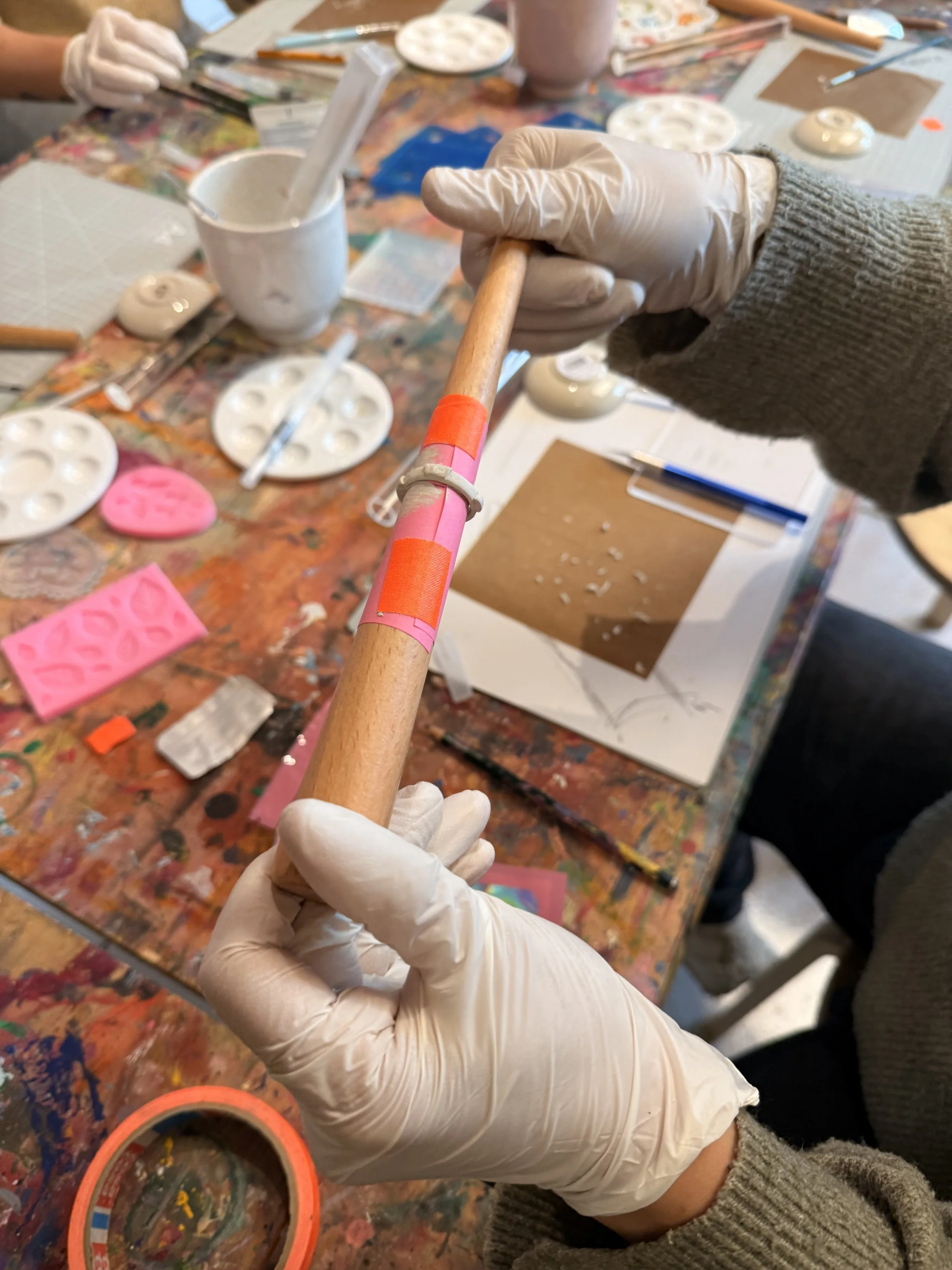 Person hält ein langes, handbemaltes Kunststück aus Holz. Im Hintergrund sind Bastelutensilien und Malpaletten auf einem bunte, schmutzige Arbeitstisch zu sehen.