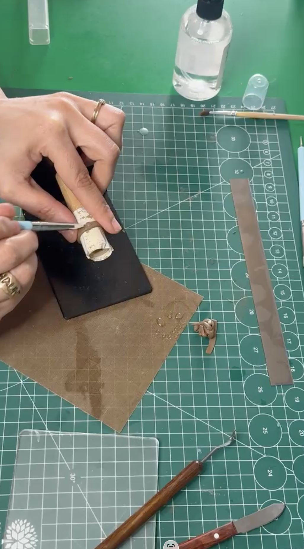 Jemand arbeitet an einem kleinen Modell aus Papier- oder Pappstücken auf einem Schneidematte, umgeben von Werkzeugen, einem Fläschchen mit Wasser und anderen Bastelmaterialien.