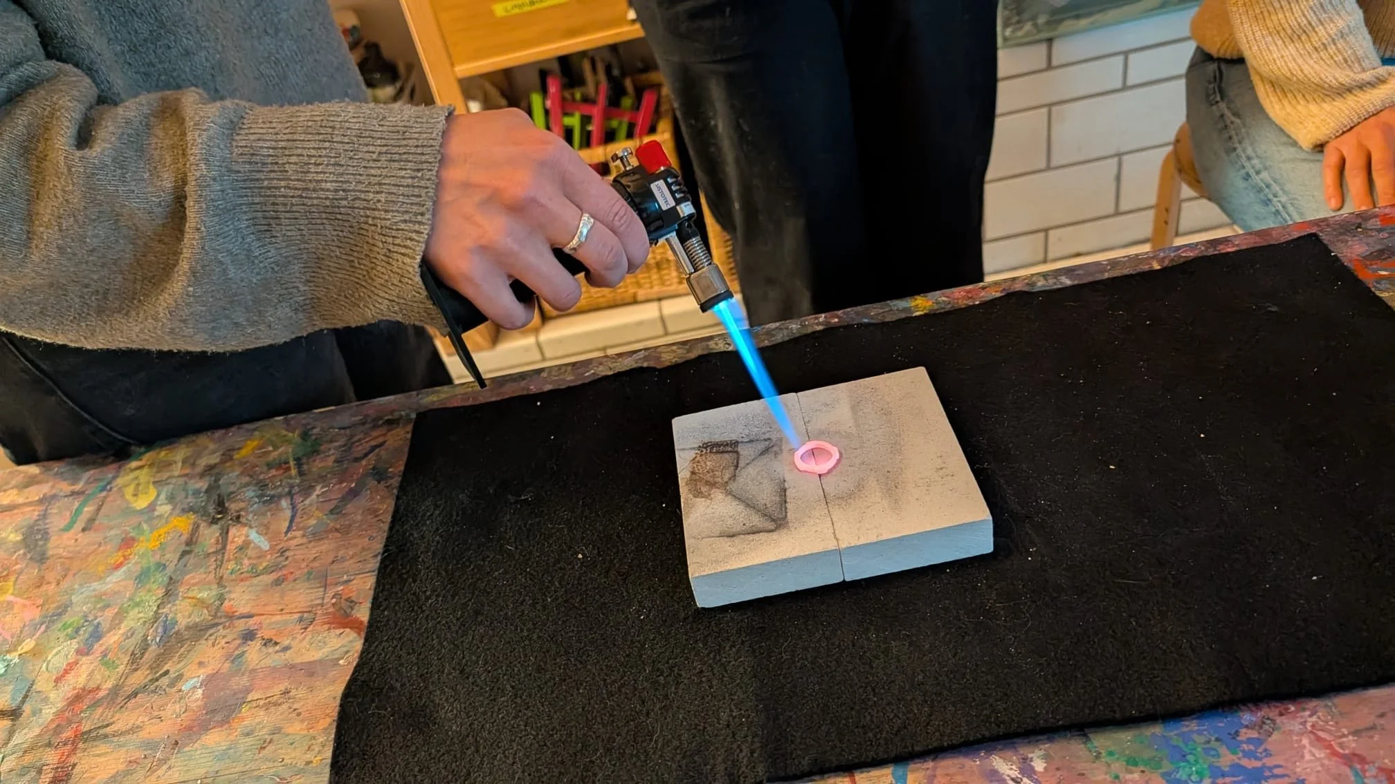 Eine Person benutzt einen Gasbrenner, um an einem runden Gegenstand zu arbeiten, der auf einem weißen Styroporblock liegt. Im Hintergrund sind Regale mit bunten Gegenständen.