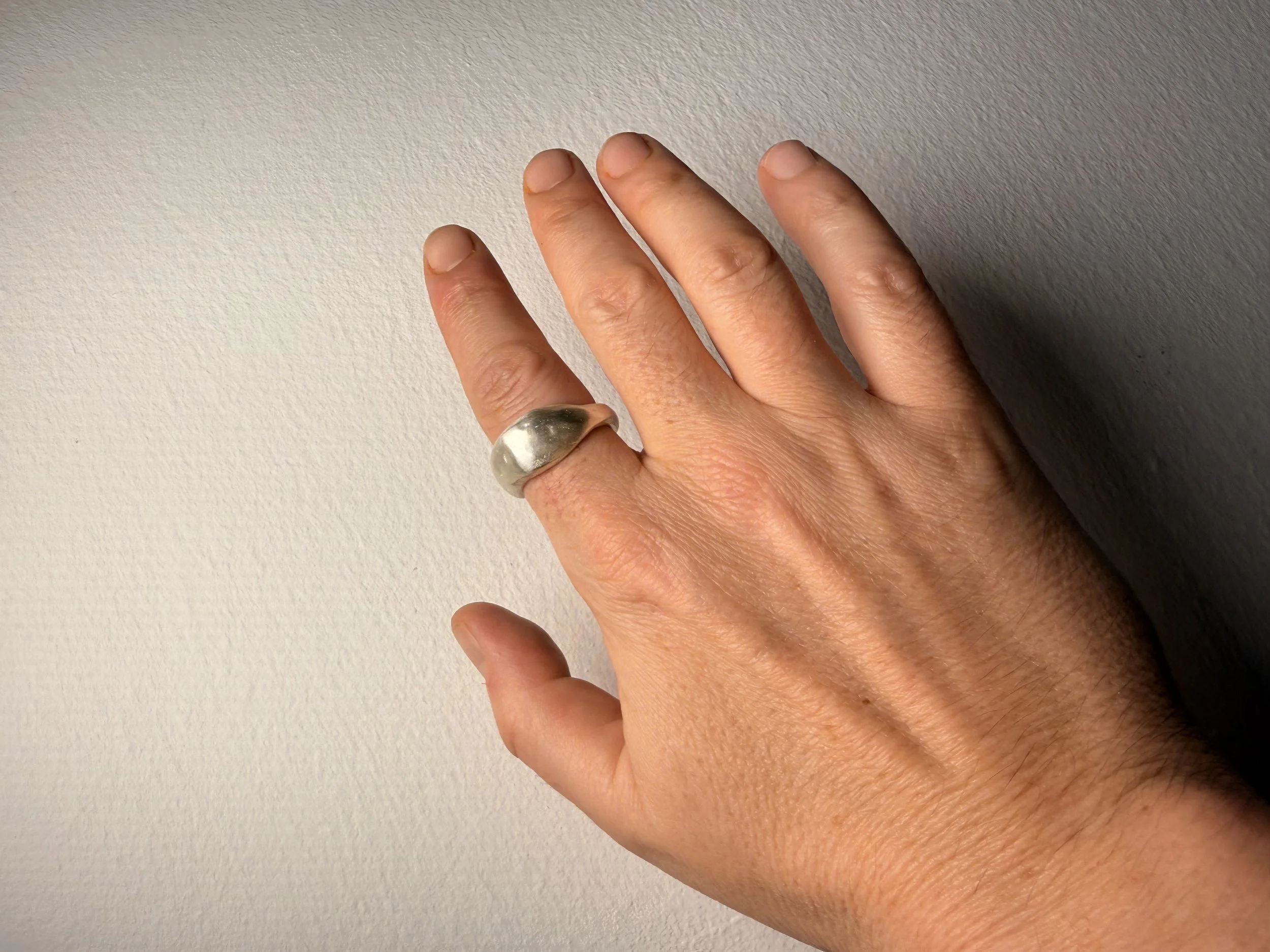 Nahaufnahme einer menschlichen Hand mit einem großen, silberfarbenen Ring am Finger. Die Hand liegt auf einer weißen Oberfläche und die Fingernägel sind unregelmäßig und gepflegt.