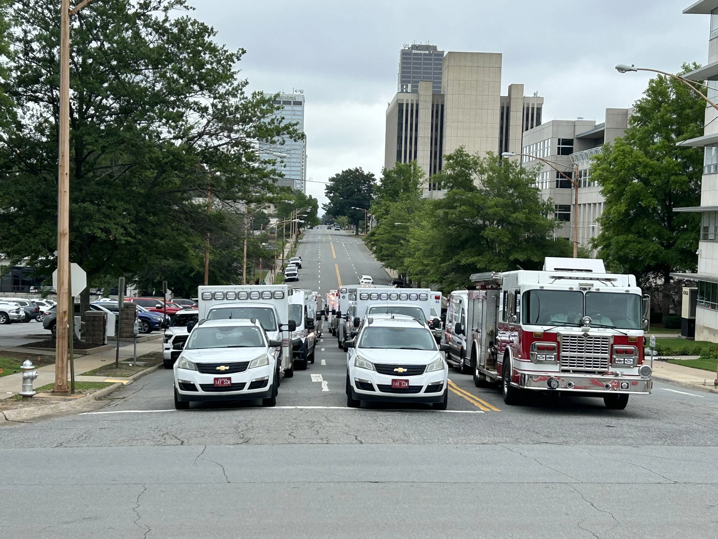 View of an urban street with several parked and moving vehicles, multiple trees lining the sides, and tall office buildings in the background on a cloudy day.