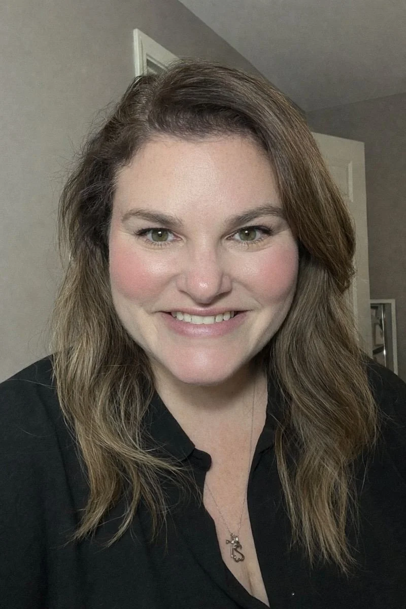 A woman with shoulder-length wavy brown hair, light skin, and green eyes smiling in a black shirt with a necklace, interior background with gray walls and a door.