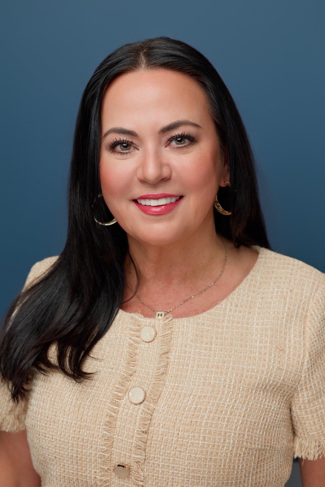 Headshot of a woman with long dark hair, wearing a beige textured top and jewelry, smiling in front of a blue background.